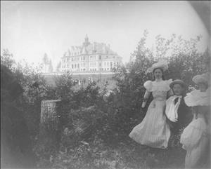 Four children, three girls and one boy, stand in a small clearing. There is a large castle like building, with turrets, towers and a pointed roof in the distance. The girls are wearing long frilly dresses with mutton chop sleeves and white hats, the boy is dressed in a sailor suit and wears a boater hat.  