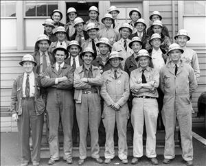 A smiling group of men stand on a set of stairs, wearing work clothing, and ties. They have their arms crossed, wearing hard hats and stand against the backdrop of a house with large windows. 