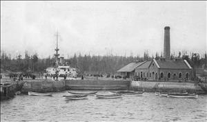 A large white boat in a small dock, surrounded by people, and a small brick building with a smoke stack. In the front are smaller boats, and calm water. 