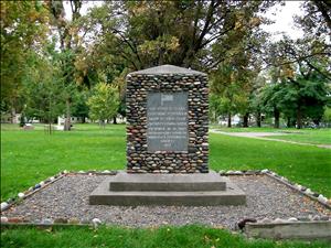 A stone marker set in a larger pebbled marker on a small pedestal. There is green grass in the background, and tall trees. 