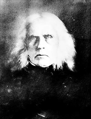 Head and shoulders portrait of an older man with white shoulder length hair, a black jacket, and wide-eyed expression.
