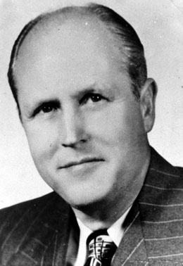 Head and shoulders portrait of a man wearing a tie, and striped jacket. He is balding, and has a slight smile on his face. 