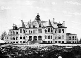 A large four story castle shaped mansion in the Queen Anne style, with three arches above the doorway, many turrets and towers. It is set back slightly on a hill. 