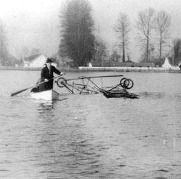 A man wearing a suit and hat canoes past an upside down plane in a lake, its wheels sticking out. He has a paddle in his right hand, and there are trees and houses in the background.