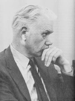 An older man with white hair and a mustache dressed in a collared shirt, suit coat and tie. He looks off to the left with a pensive expression, his hand to his lips. 