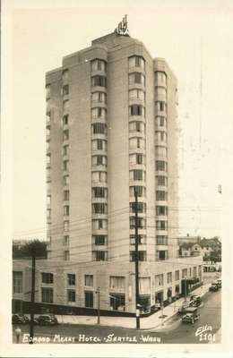 A fourteen story raised tower hotel with many windows, larger at the base than the top. There is a small entryway with a covered roof, and many antique cars parked on the street. Houses and trees are in the background. 