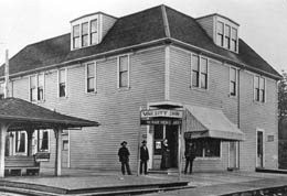 Four people stand outside a large wooden building with two dormer windows. There is a sign above the door, the men wear hats, and the woman is wearing a dress. 